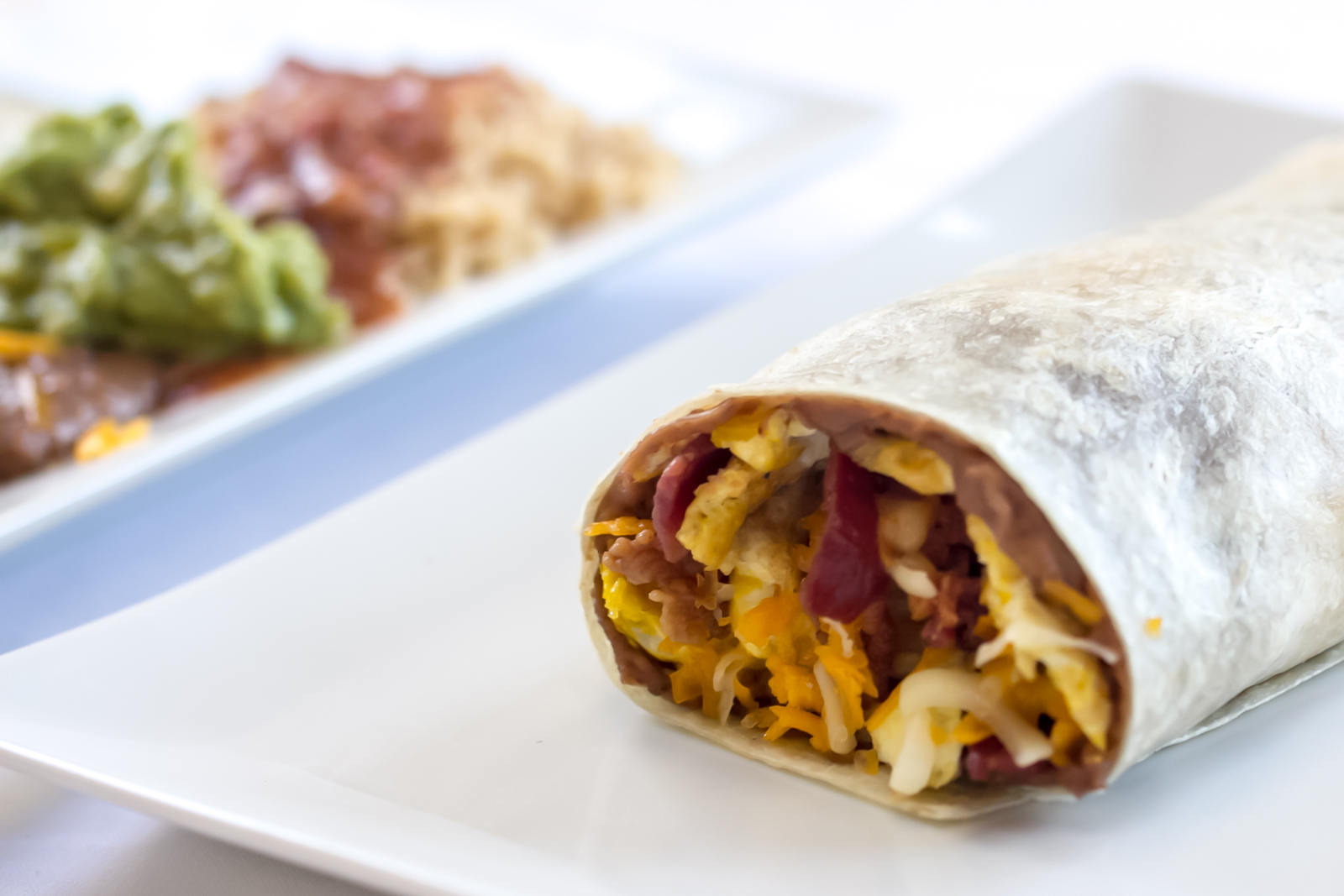 Close-up of a rolled breakfast burrito with a plate of guacamole, rice and beans blurred behind it.
