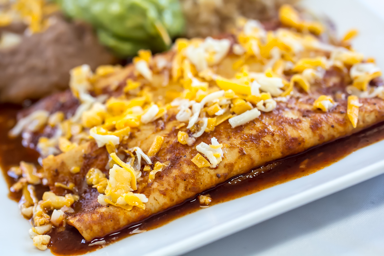 Two enchiladas covered in red chili sauce and shredded cheese, plated with refried beans, rice and guacamole.