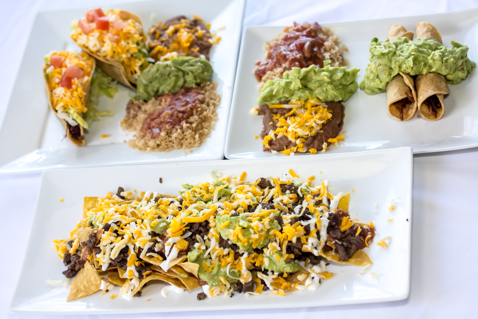 Three white plates holding hard tacos, taquitos with guacamole, and a long plate of nachos.