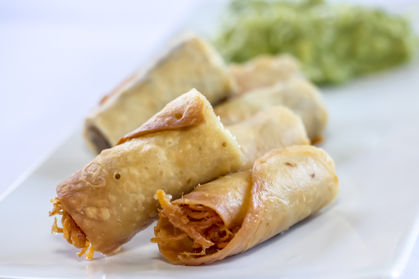 Four golden taquitos on a white plate with guacamole behind them.
